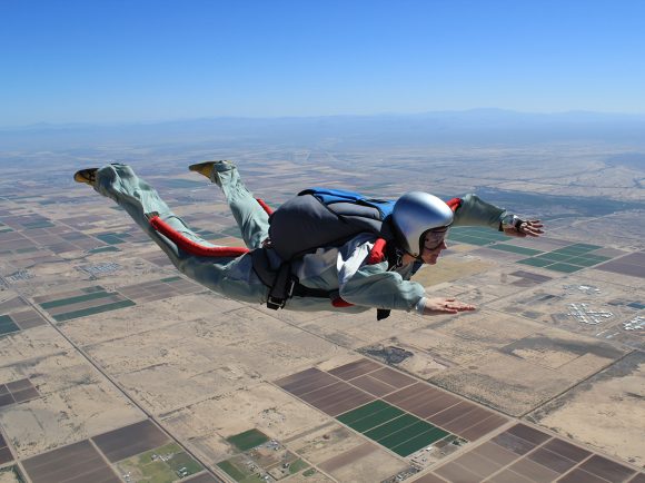 Erin Bonilla skydiving in a silver jumpsuit and helmet over Skydive Arizona